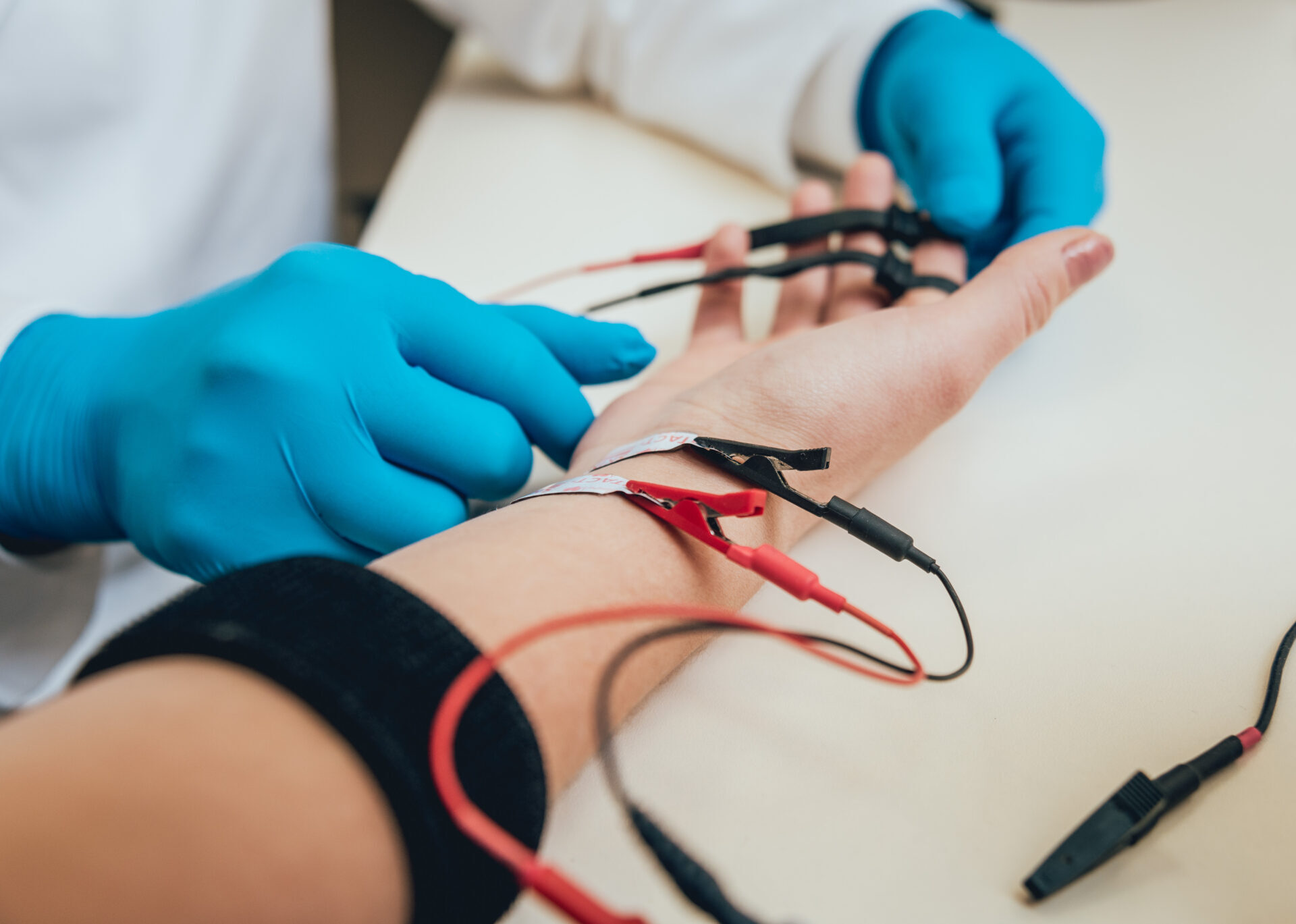 Ārsts ar zilu cimdu pievieno elektroda vadiņus pie pacienta rokas. Redzami sarkani un melni vadi.