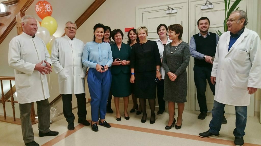 Rīgas 1. slimnīcas darbinieki svin jubileju, stāvot kopā pie kāpnēm ar baloniem un smaidiem.