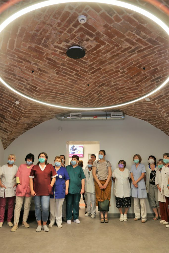 Rīgas 1. slimnīcas darbinieki grupā, tērpti medicīniskajās drēbēs un maskās, stāv telpā ar ķieģeļu griestiem.