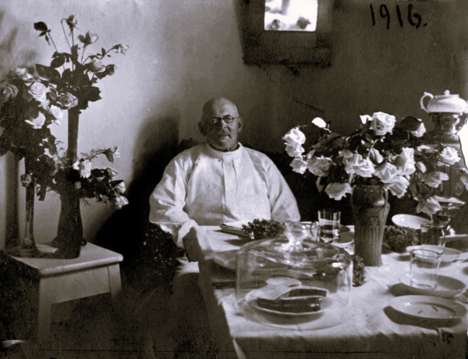 Vīrietis baltā apģērbā sēž pie galda ar ziediem un ēdienu, fiksēts 1916. gadā.