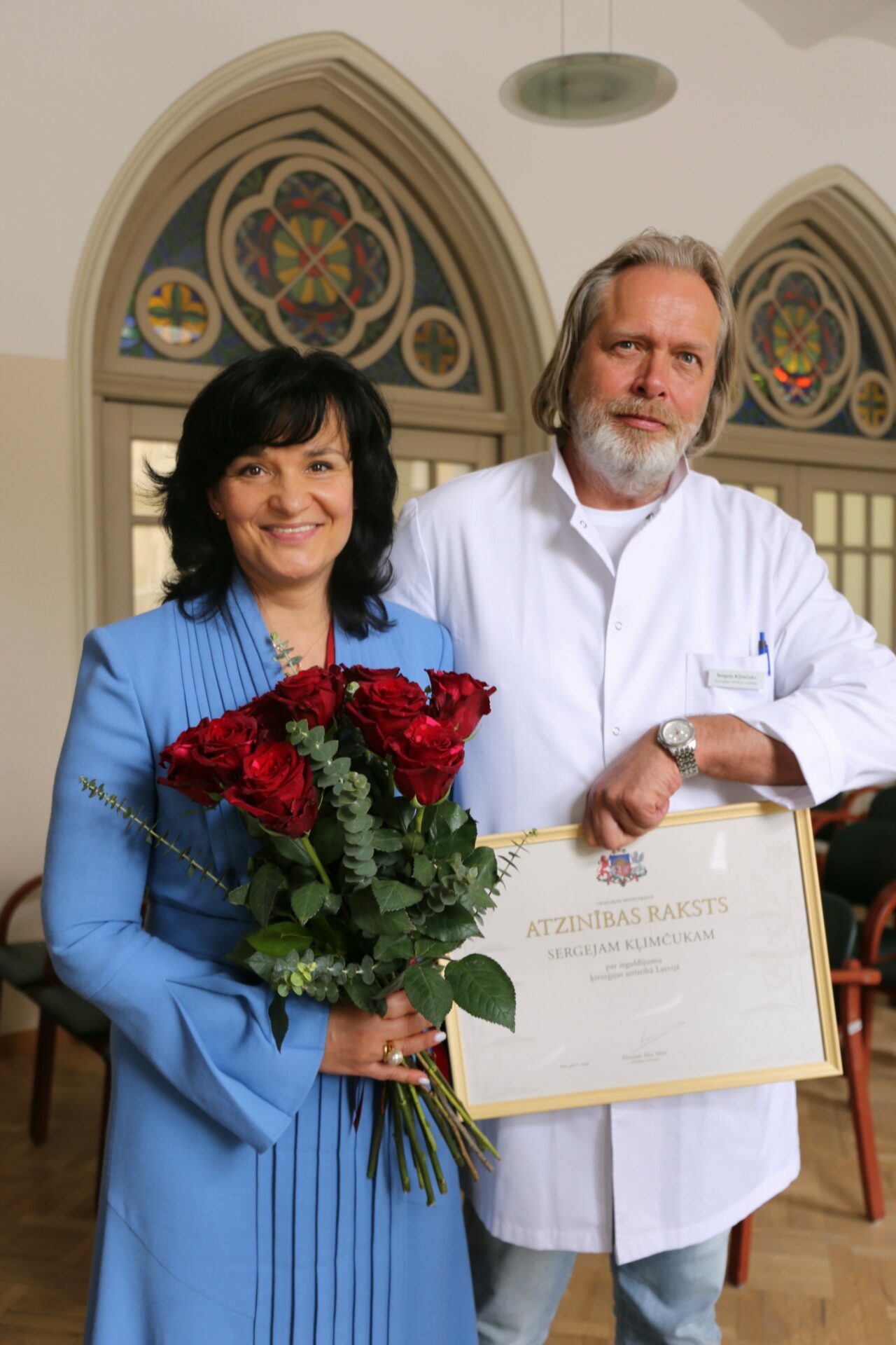 Divi cilvēki pozē ar ziediem un atzinības rakstu, uz kura rakstīts 