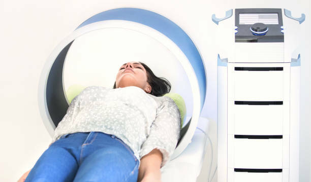 Sieviete guļ uz gultas, izmantojot medicīnisko iekārtu Rīgas 1. slimnīcā. Redzama iekārta un kontroles panelis.