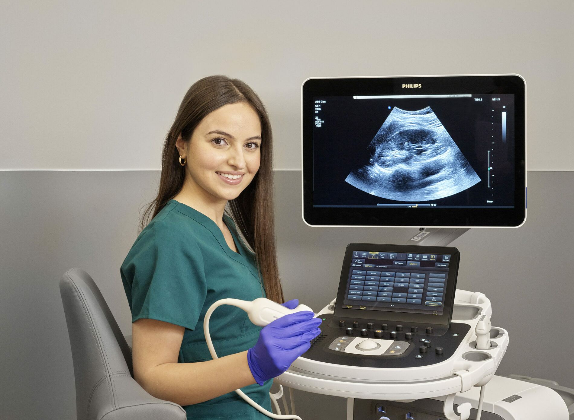 Jauna sieviete zaļā medicīnas tērpā ar ultraskaņas aparātu un ekrānu, kurā redzams ultraskaņas attēls.
