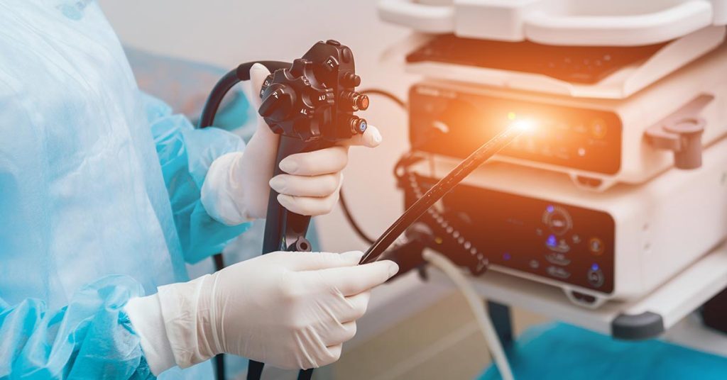 Ārsts tur endoskopu, gatavojoties procedūrai. Fons ar medicīnas iekārtām Rīgas 1. slimnīcā.