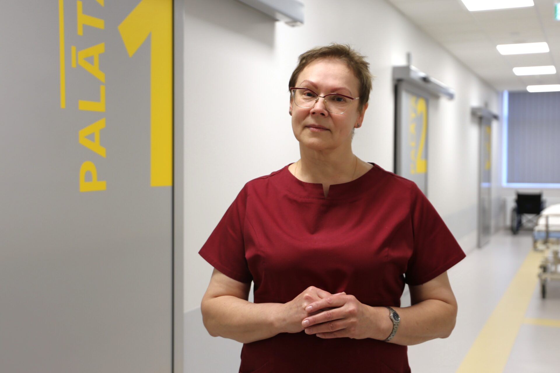 Sieviete sarkanā medicīnas tērpā stāv pie durvīm ar uzrakstu "PALĀTA 1" Rīgas 1. slimnīcā.