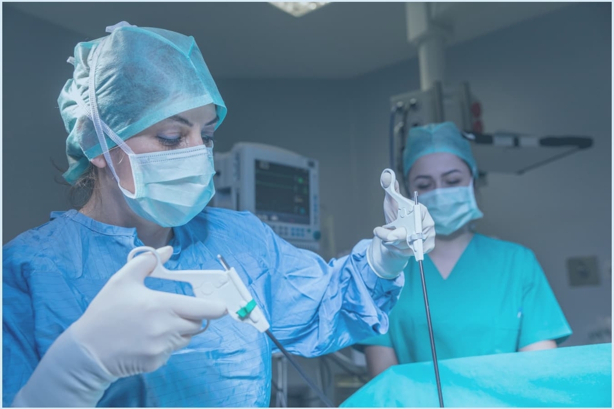 Divas medicīnas darbinieces veic operāciju, tērptas ķirurģiskajā apģērbā un maskās. Redzami instrumenti.