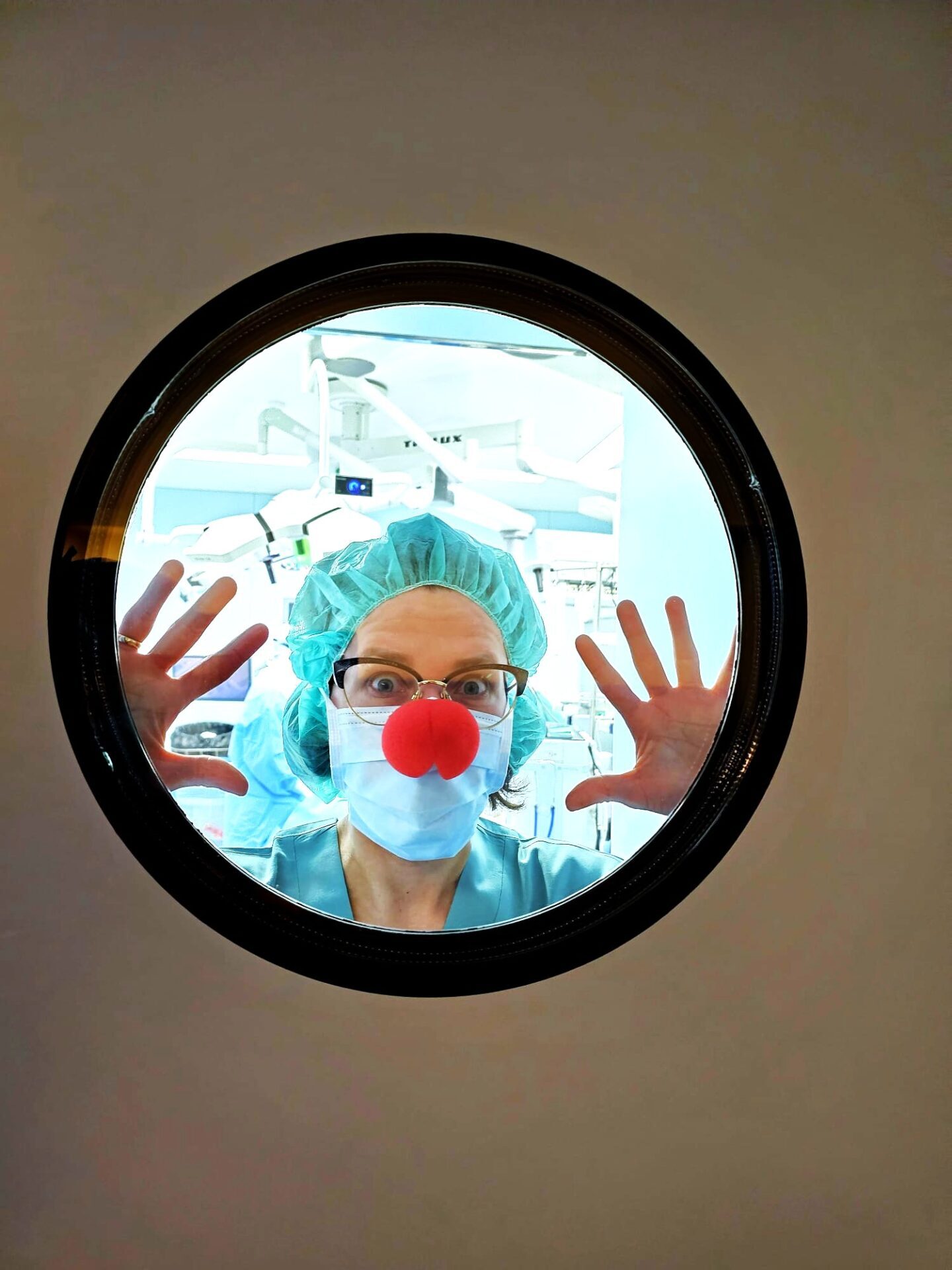 Sieviete ar ķirurģisko masku un sarkanu degunu skatās caur apaļu logu, tērpta medicīniskajā apģērbā.