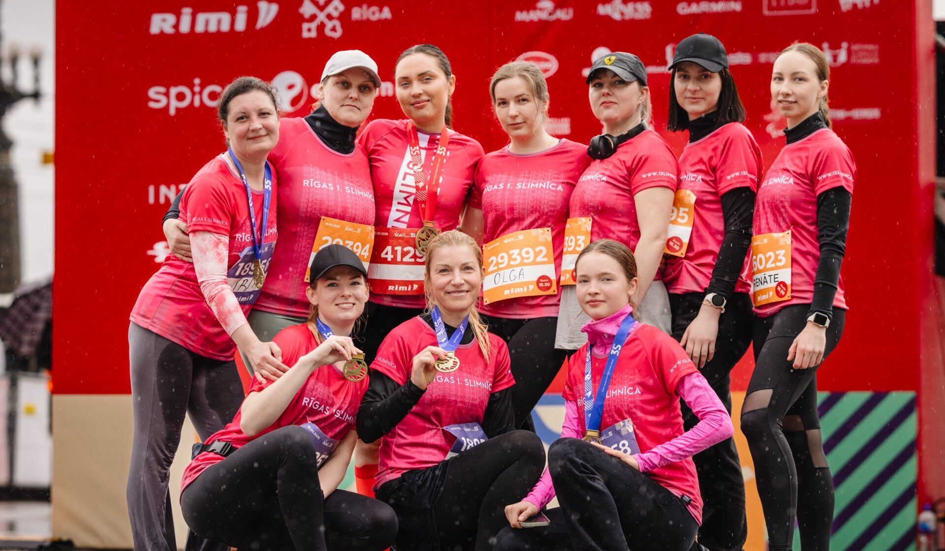 Desmit sievietes ar medaļām, tērptas rozā T-kreklos ar uzrakstu "Rīgas 1. slimnīca", pozē grupā.