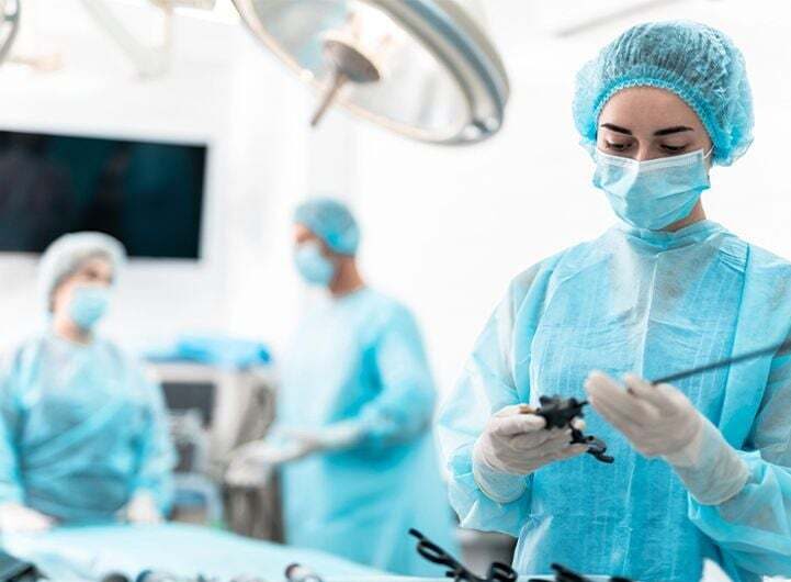 Ķirurgs operāciju zālē, tērpies zilos aizsargtērpos, tur instrumentus, blakus citi medicīnas darbinieki.