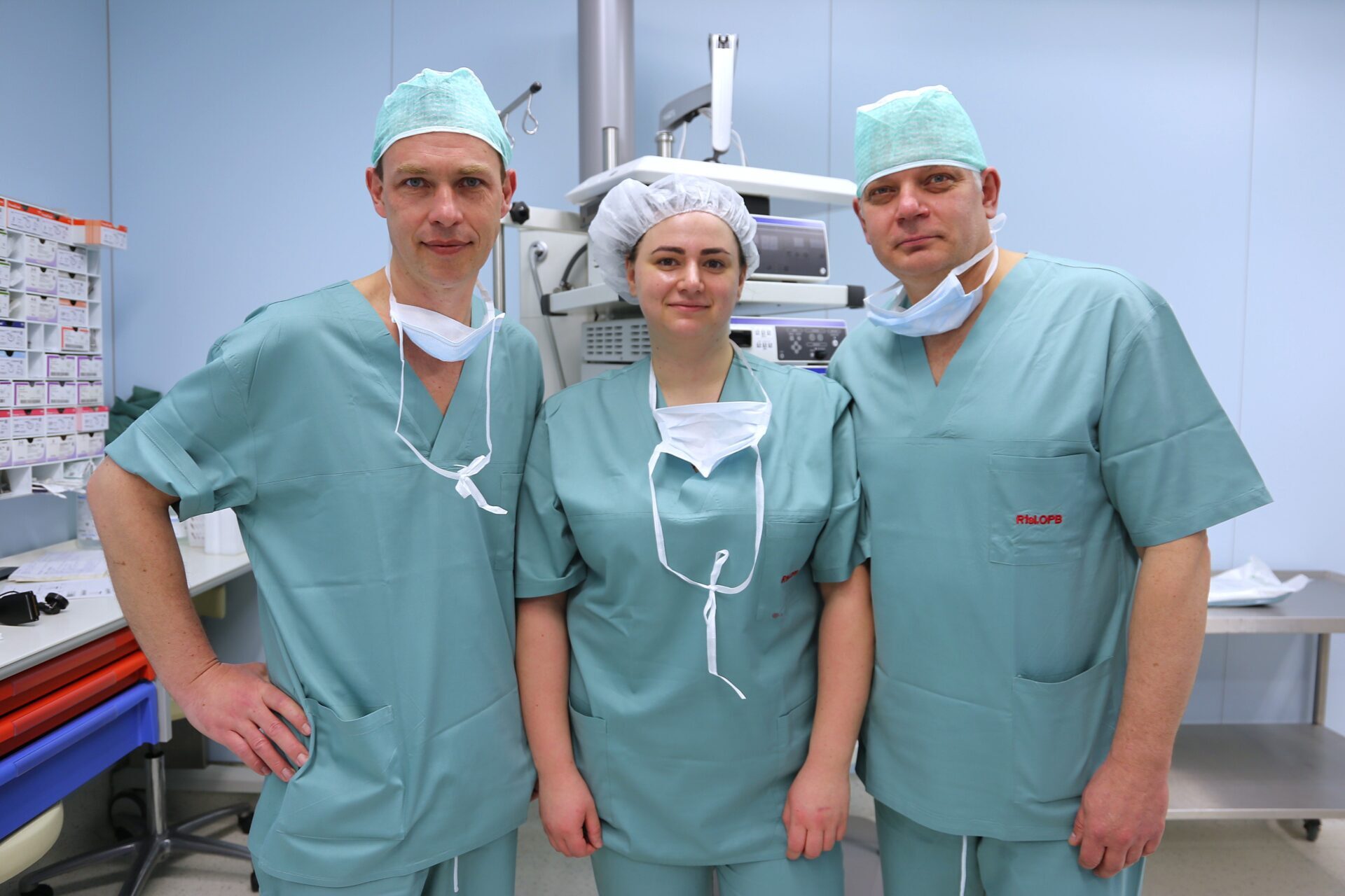 Trīs mediķi zaļās operāciju drēbēs stāv slimnīcas telpā, fons ar medicīnas iekārtām un instrumentiem.