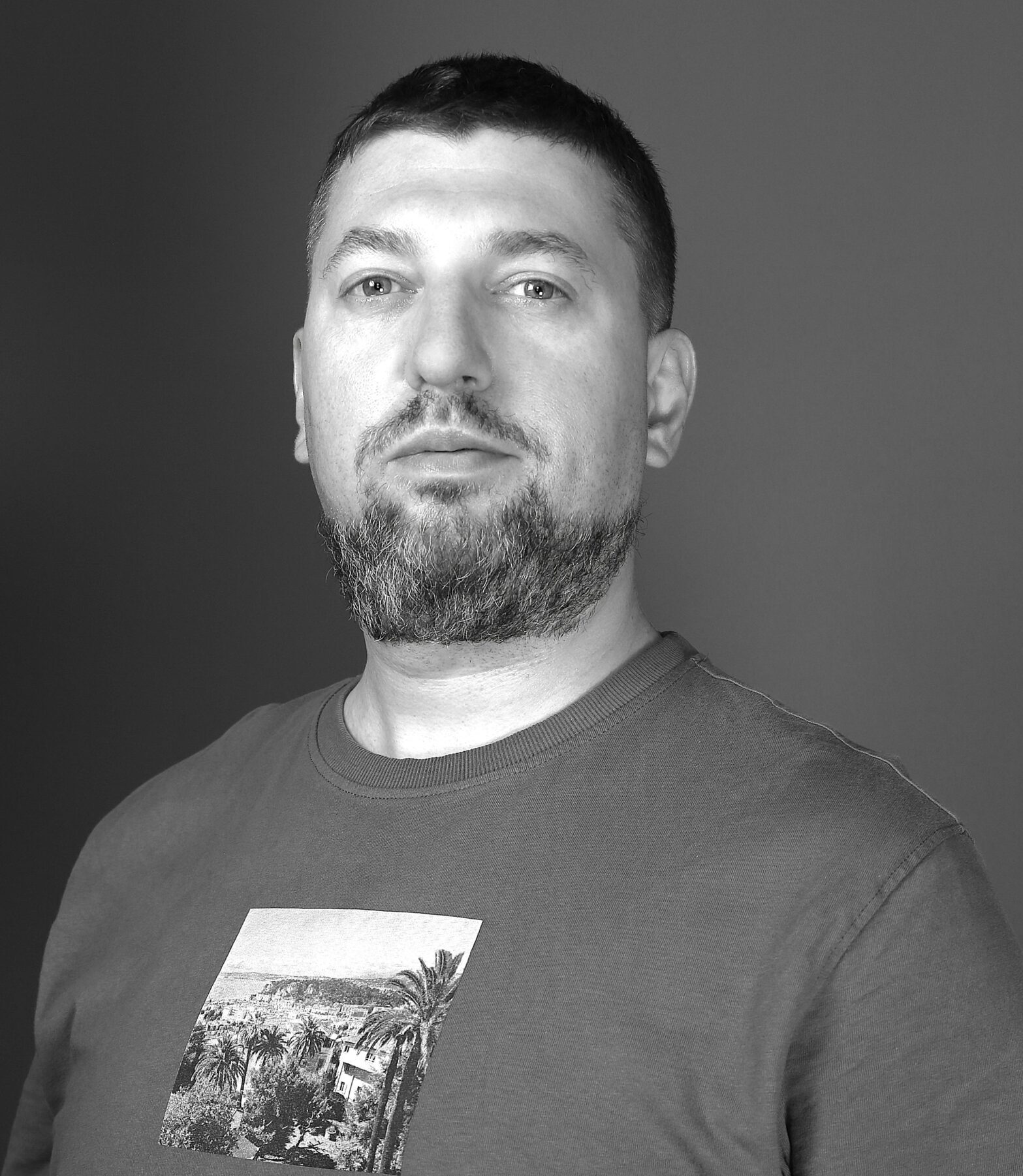 Vīrietis ar bārdu, pelēkā kreklā ar attēlu, skatās tieši uz kameru. Fons ir pelēks.