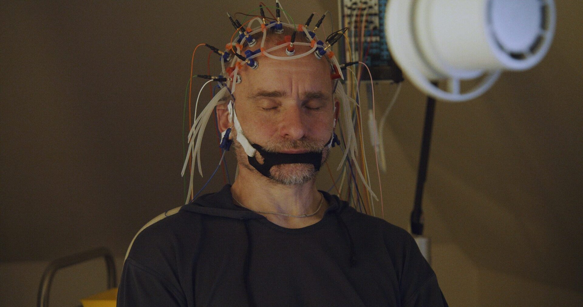Vīrietis ar elektrodiem uz galvas, veicot medicīnisku procedūru Rīgas 1. slimnīcā. Acis aizvērtas.