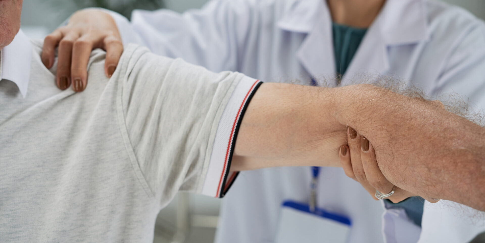 Ārsts palīdz pacientam, turēdams viņa roku, Rīgas 1. slimnīcā. Redzama medicīniska apģērba detaļa.