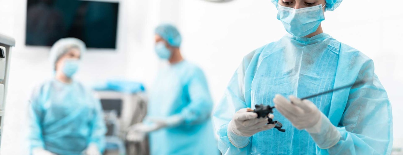 Ārsts operācijas telpā, tērpies zilos medicīniskos tērpos, tur instrumentu. Fons neskaidrs, redzami citi mediķi.