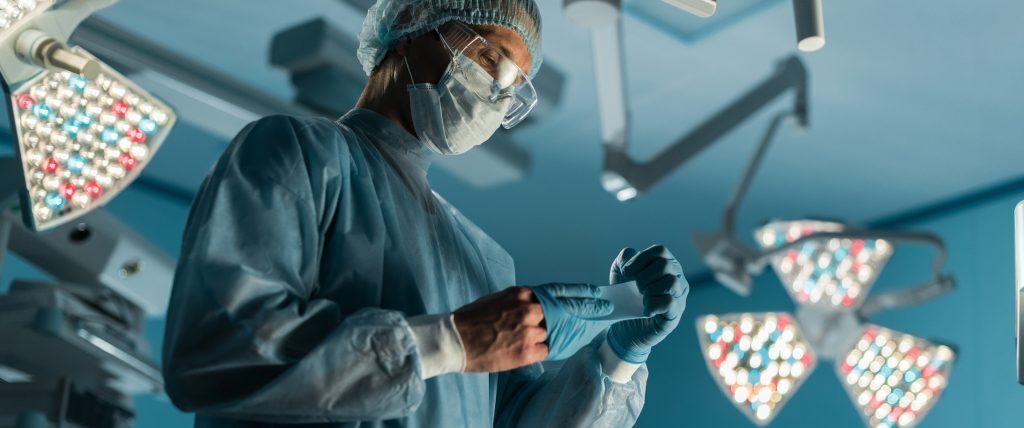 Ārsts operāciju zālē uzliek cimdus, sagatavojoties procedūrai. Apgaismojums ir spilgts un moderns.