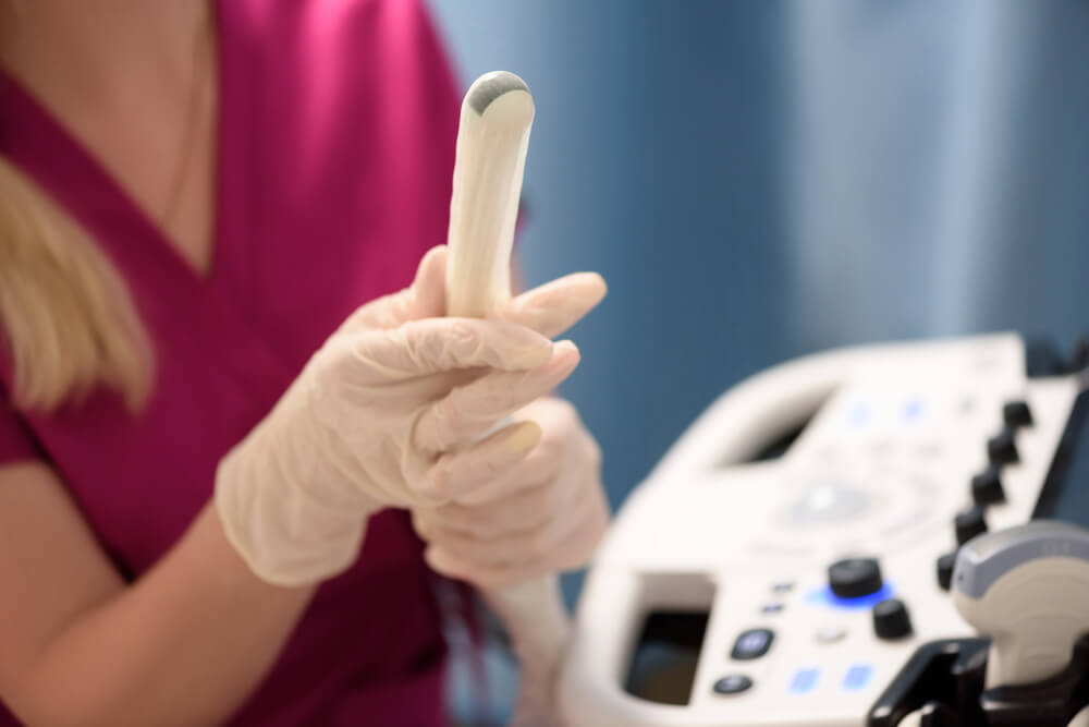 Mediķis ar cimdiem tur ultraskaņas ierīces daļu, fons ir medicīniska aprīkojuma panelis.