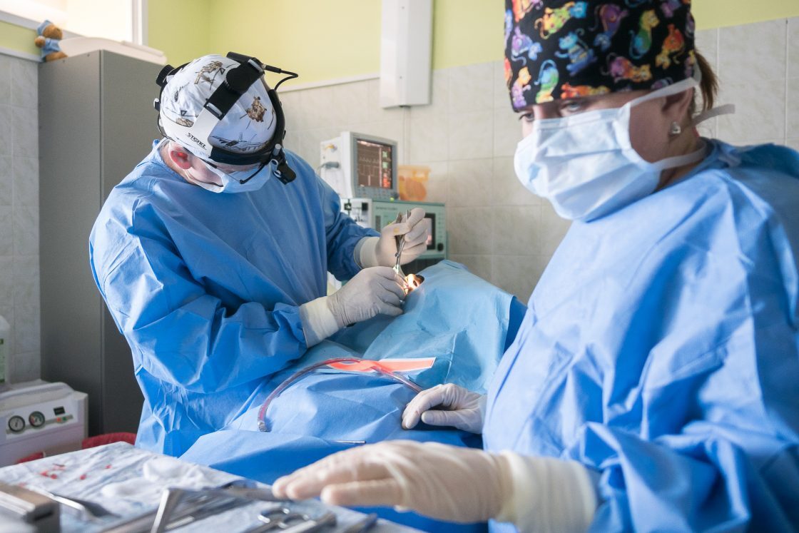 Divi ķirurgi veic operāciju, valkājot zilu ķirurģisko apģērbu un maskas. Redzami medicīnas instrumenti.