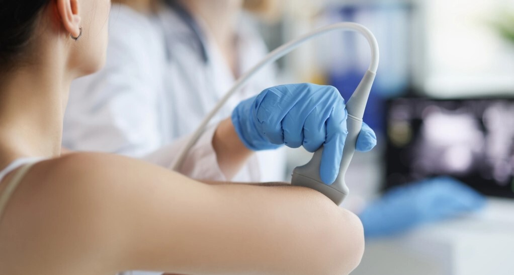 Ārsts veic ultraskaņas izmeklēšanu uz pacienta rokas. Redzami medicīnas instrumenti un zils cimds.