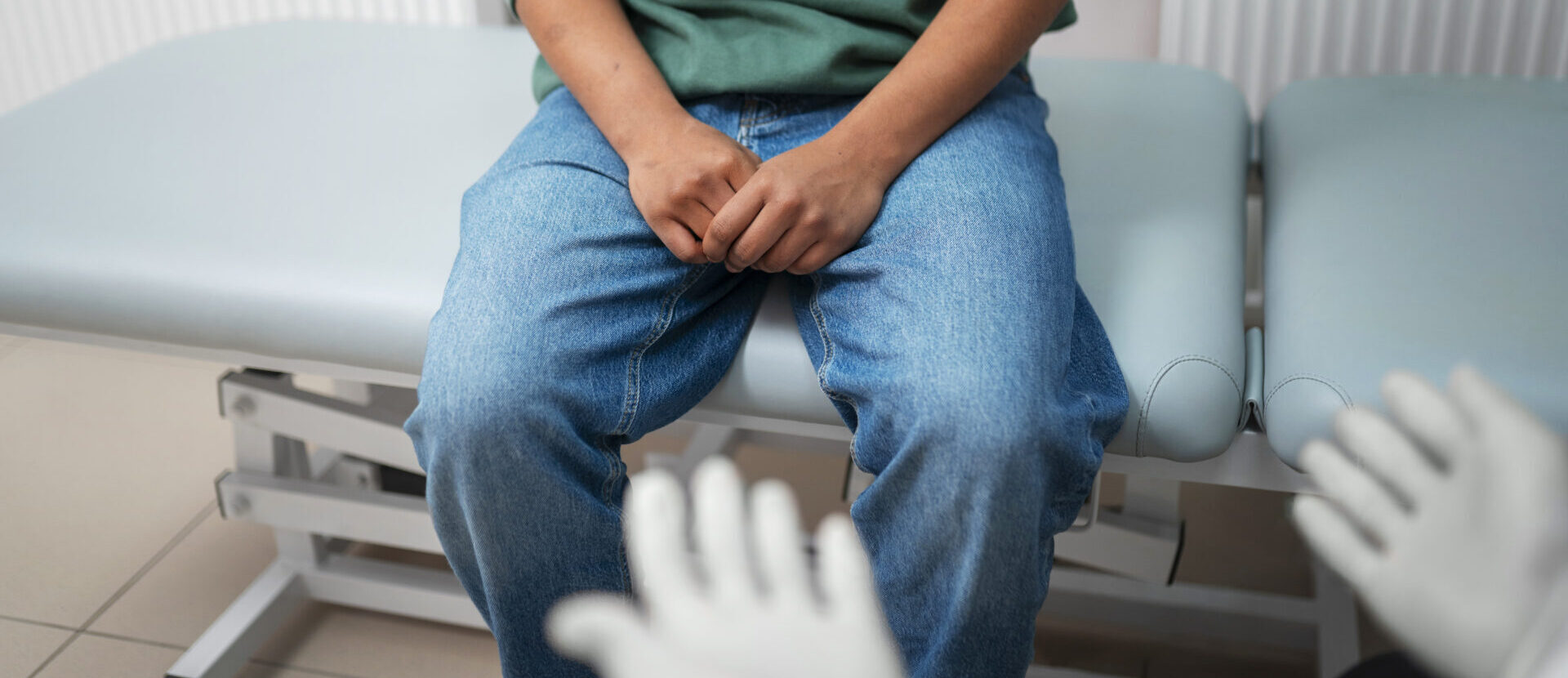Bērns sēž uz medicīniskā galda, rokas saliktas uz ceļiem, redzamas baltas medicīniskās cimdi.