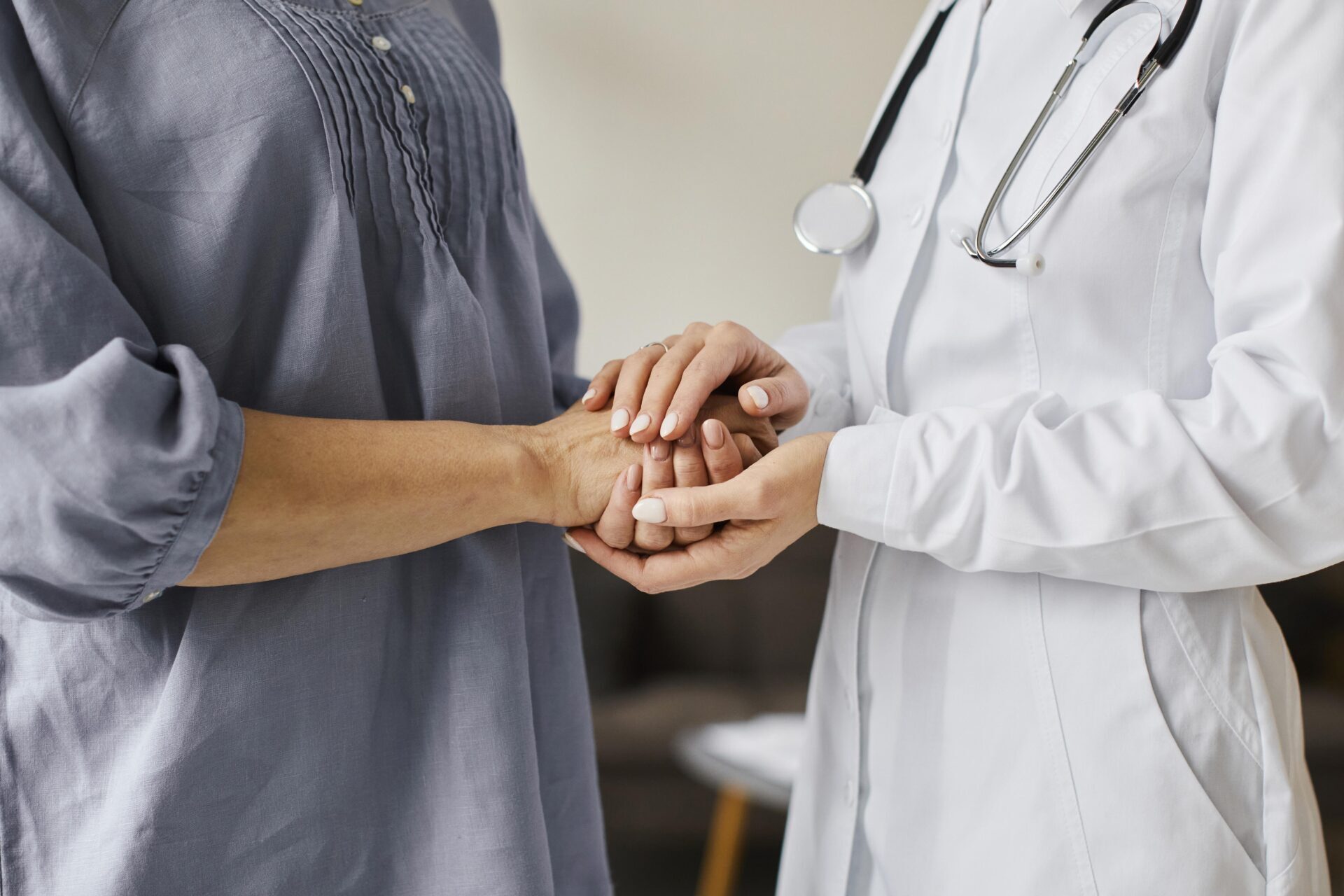 Ārsts tur pacienta roku, sniedzot atbalstu. Attēls ilustrē medicīnisko aprūpi Rīgas 1. slimnīcā.