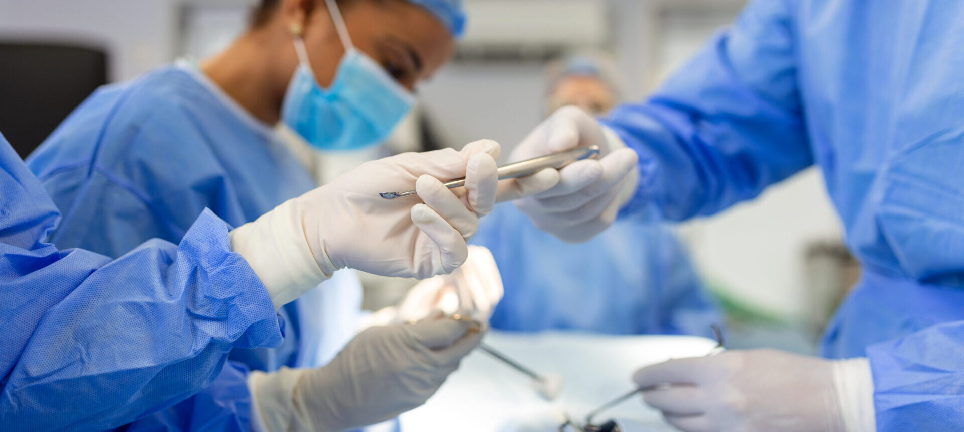 Ārsti veic operāciju, tērpti zilos medicīniskos apģērbos un cimdos, izmantojot instrumentus.