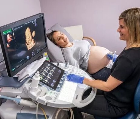 Grūtniece guļ uz gultas, kamēr ārste veic ultrasonogrāfiju ar ekrānu, kur redzams augļa attēls.