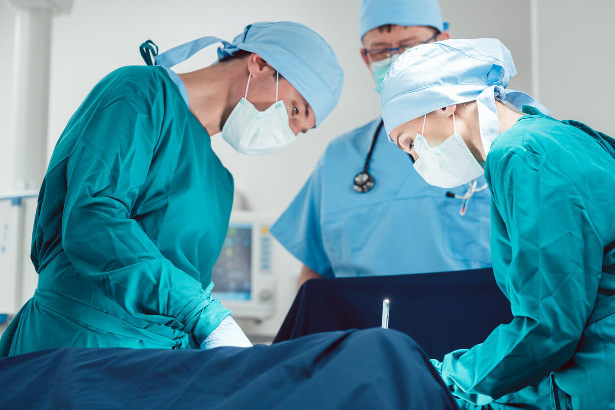 Trīs ķirurgi operācijas laikā, tērpti zaļās un zilās medicīnas drēbēs un maskās. Rīgas 1. slimnīca.