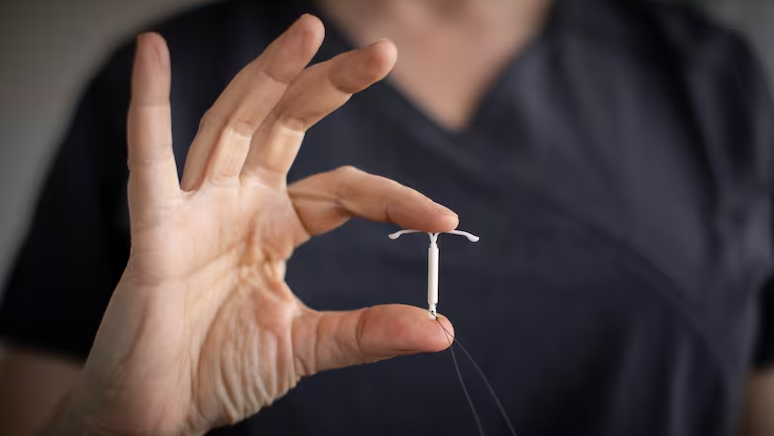 Rokā tur IUD (intrauterīnā ierīce), kas ir kontracepcijas metode. Fons ir medicīnisks apģērbs.