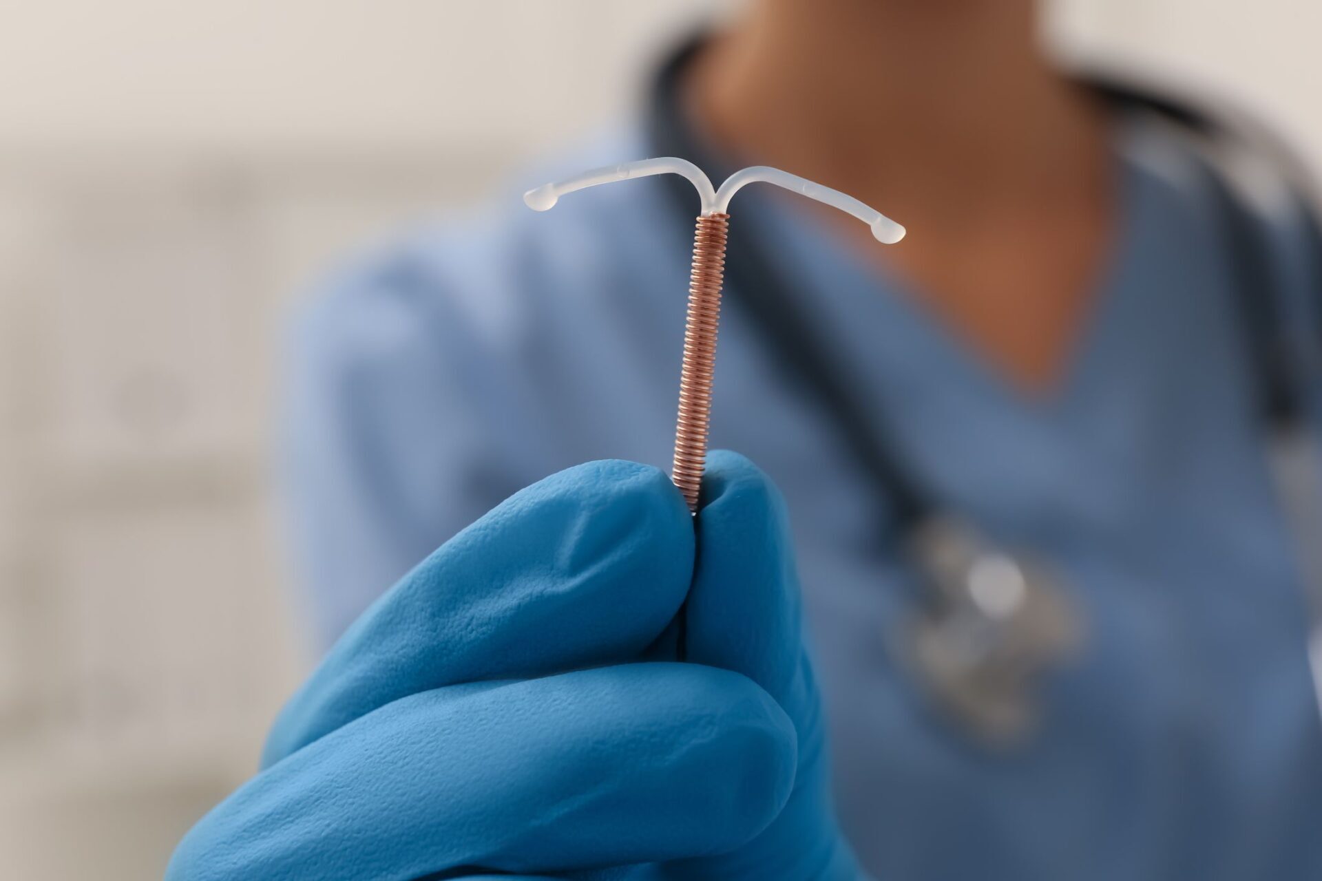 Ārsts tur IUD kontracepcijas līdzekli, valkājot zilu cimdu. Fons izplūdis, redzams medicīniskais aprīkojums.