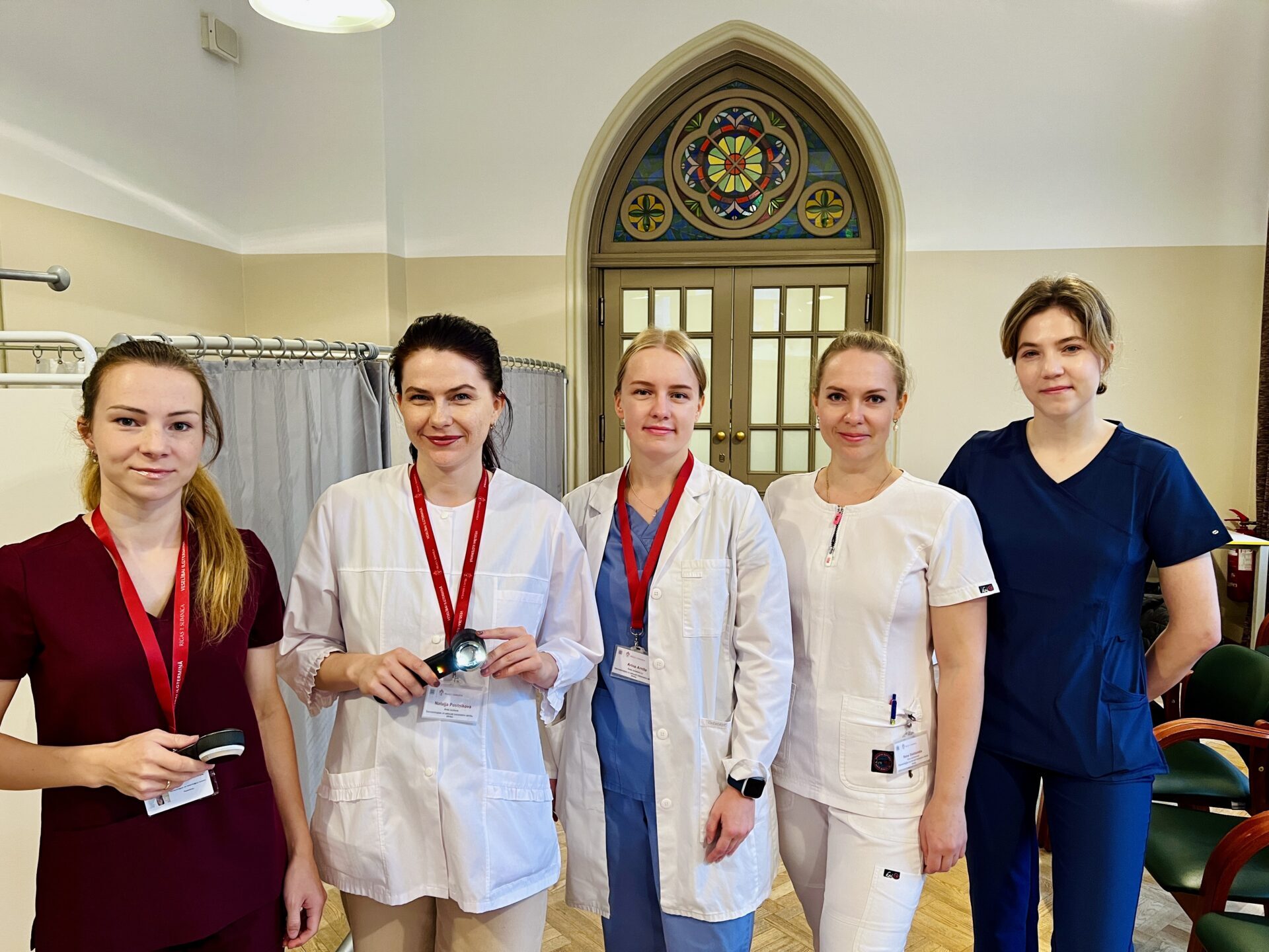 Pieci medicīnas darbinieki Rīgas 1. slimnīcā, tērpti darba apģērbā un ar identifikācijas karšu kakla siksnām.