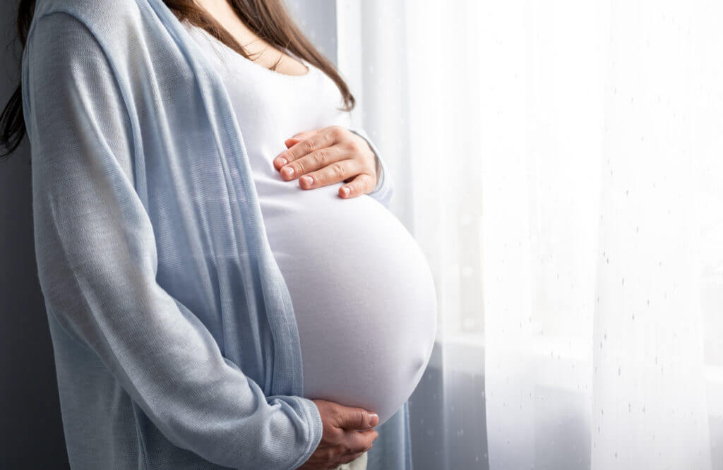 Grūtniece stāv pie loga, turot roku uz vēdera, apģērbtā gaišā žaketē. Gaisma ieplūst caur aizkariem.