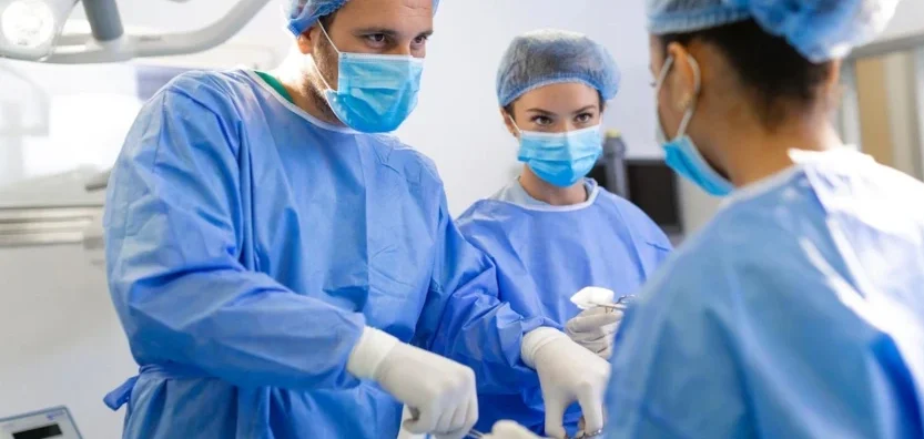 Trīs medicīnas darbinieki operāciju zālē, valkā zilu apģērbu un sejas maskas.