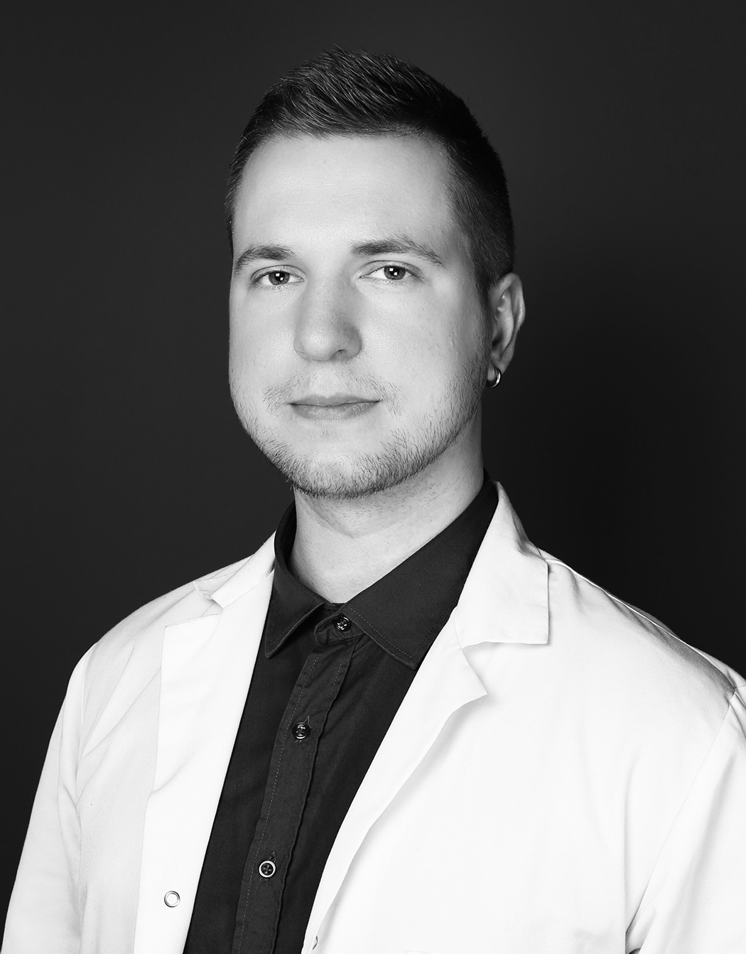 Jauns ārsts baltā mētelī, melnā kreklā, ar īsiem matiem un auskaru, Rīgas 1. slimnīcas kontekstā.
