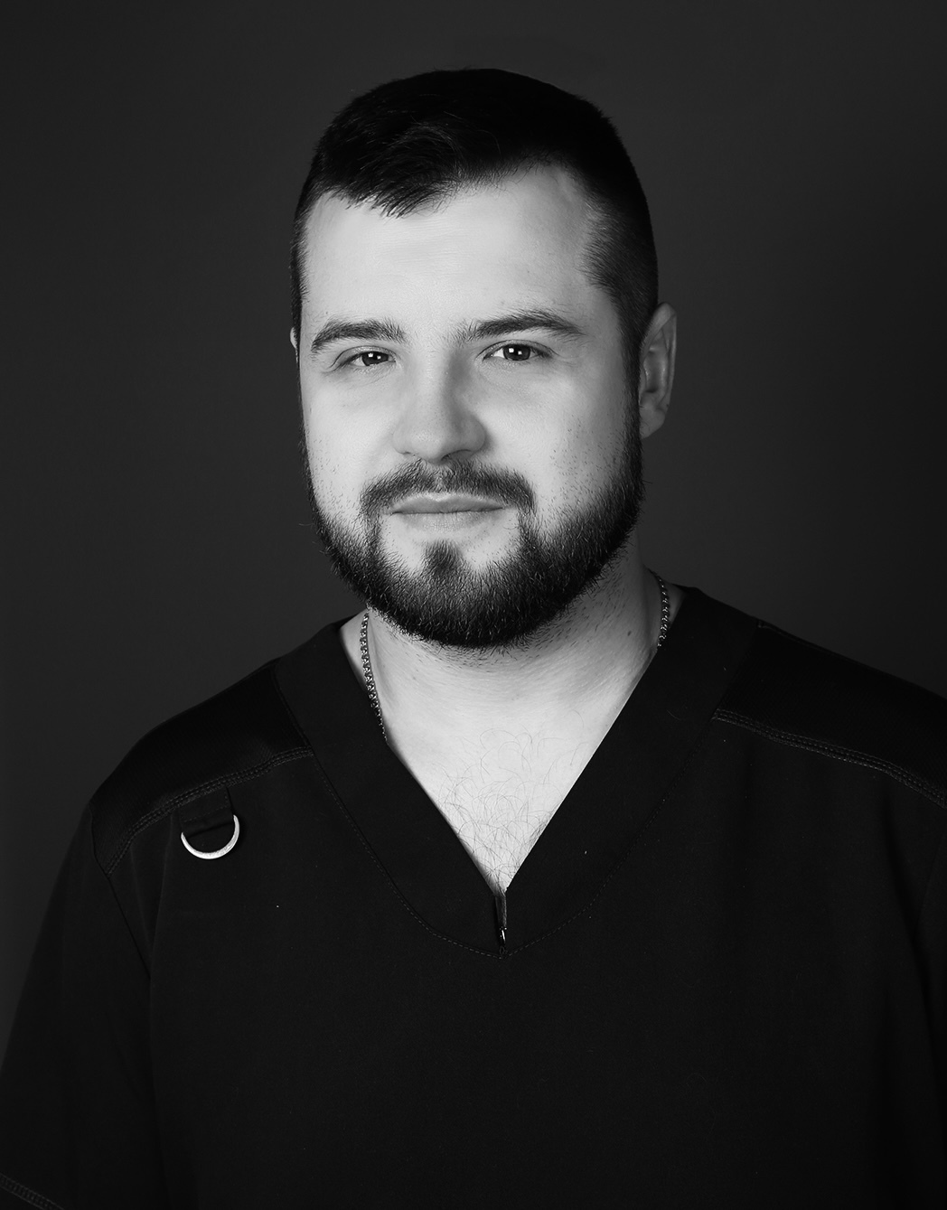 Vīrietis ar bārdu, melnā medicīnas apģērbā, skatās uz kameru. Rīgas 1. slimnīcas darbinieks.