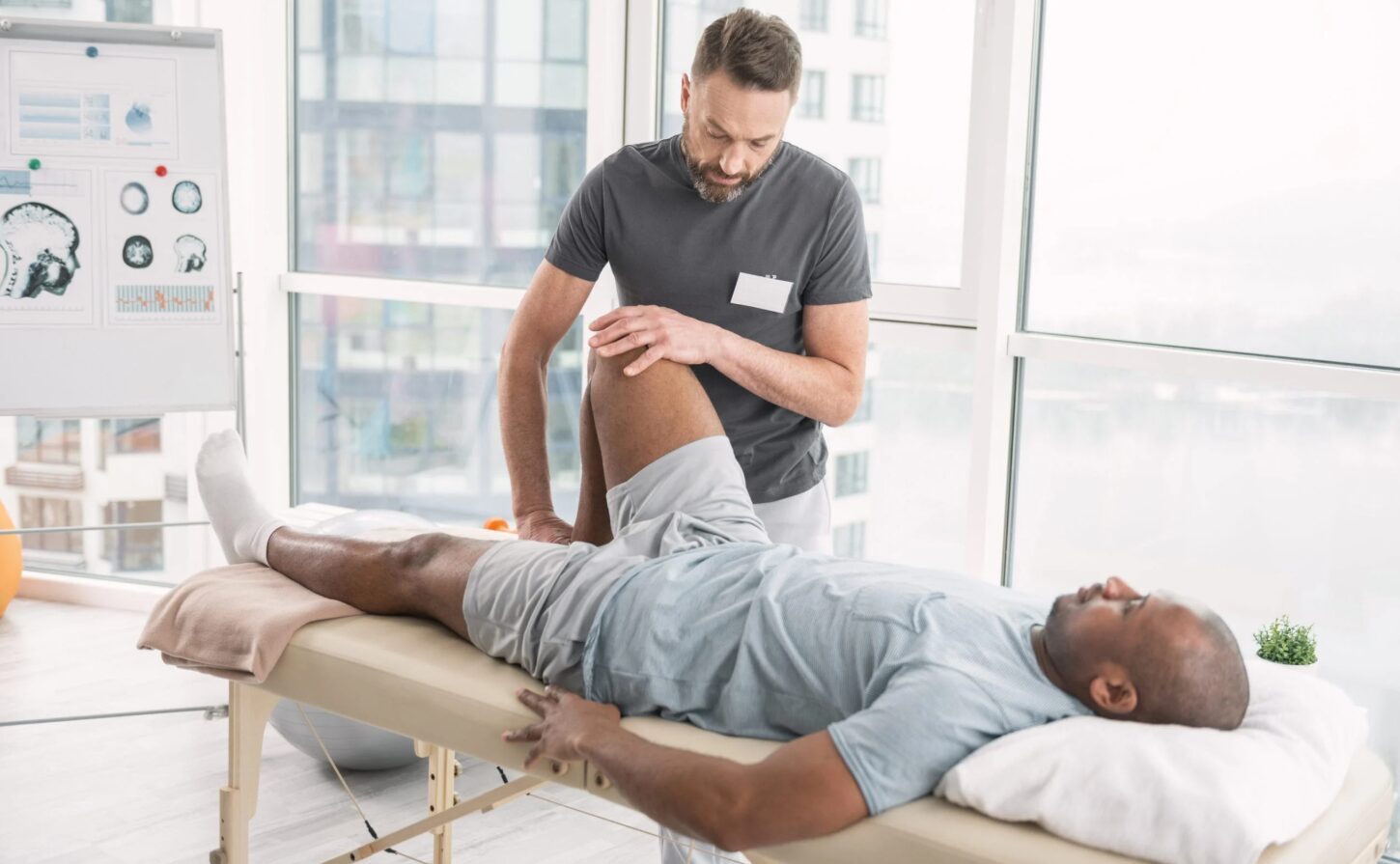 Fizioterapeits veic ārstēšanu pacientam uz masāžas galda Rīgas 1. slimnīcā. Aiz sienas ir medicīnas attēli.