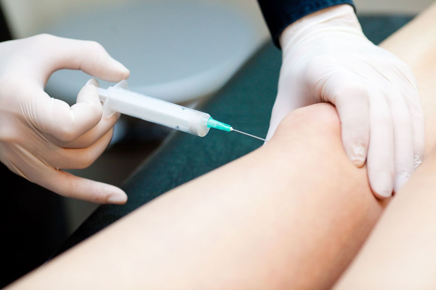 Medicīnas darbinieks veic injekciju pacientam, turot šļirci un pieskaroties pacienta kājai.