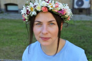 Jauna sieviete ar ziedu vainagu, sēžot zālē, ar gaišām acīm un zilu kreklu. Fons ir zaļš un saulains.