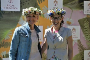 Divas sievietes ar ziedu vainagiem stāv pie Rīgas 1. slimnīcas fona, smaidot un pozējot fotogrāfijai.