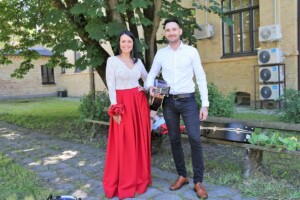 Jauna pāris ar instrumentu, stāv dārzā pie Rīgas 1. slimnīcas, sieviete sarkanā kleitā, vīrietis ar ģitāru.