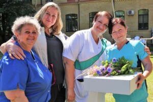 Četras sievietes, no kurām divas medicīnas darbinieces, pozē ar ziedu pušķi Rīgas 1. slimnīcas pagalmā.