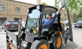 Vīrietis sēž traktortehnikā ar atvērtām durvīm, fons ar automašīnām un Rīgas 1. slimnīcas ēku.