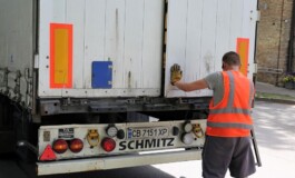 Vīrietis oranžā vestē atver kravas automašīnas aizmuguri. Redzams uzraksts "SCHMITZ" un numurs "CB 7151 XP".