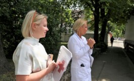 Divas sievietes stāv uz ielas, viena tērpta baltā medicīnas tērpā, otra ar dokumentiem rokās. Rīgas 1. slimnīca.