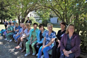 Attēlā redzama Rīgas 1. slimnīcas medicīnas personāla grupa, sēžot zem koka.