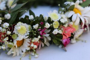 Ziedu vainags ar margrietiņām, rozēm un citiem ziediem, novietots uz balta fona.