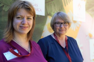 Divas sievietes Rīgas 1. slimnīcas fona priekšplānā, smaidīgas, tērptas medicīniskās drēbēs.