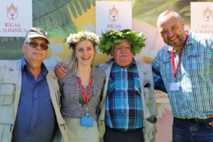 Četri cilvēki pozē pie Rīgas 1. slimnīcas fona, valkājot ziedu un lapu vainagus.