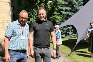 Divi vīrieši stāv saulē pie Rīgas 1. slimnīcas, fonā redzami citi cilvēki un telts.