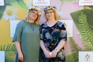 Divas sievietes ar ziedu vainagiem stāv pie Rīgas 1. slimnīcas fona ar ziediem un lapām.