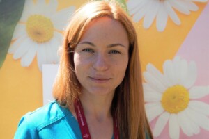 Sieviete ar sarkaniem matiem, zilu kreklu un Rīgas 1. slimnīcas identifikācijas karti, fons ar margrietiņām.